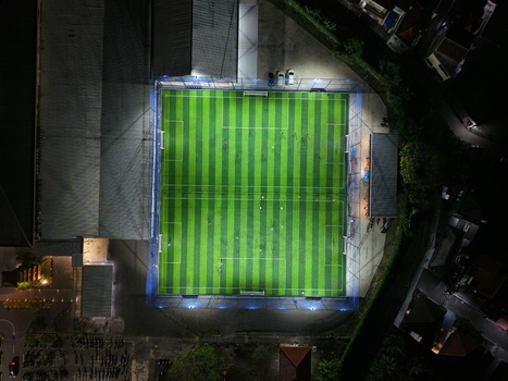 Aerial view of Bravo Arena soccer field at night illuminated by bright floodlights showing the green artificial turf and surrounding urban area in Kecamatan Denpasar Selatan Bali Indonesia.