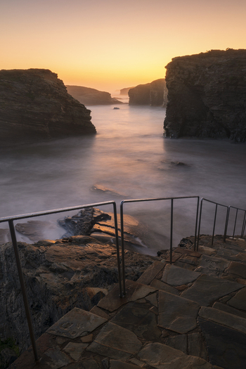 View of Praia das Catedrais with stone steps metal railings and dramatic rock formations during a golden sunrise over misty waters in Lugo Lugo Spain. View of Praia das Catedrais with stone steps metal railings and dramatic rock formations during a golden sunrise over misty waters in Lugo Lugo Spain.