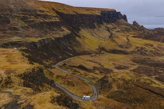 Aerial view of a white campervan driving along a winding road through the rugged cliffs and golden slopes of The Quiraing Isle of Skye Scotland United Kingdom. Aerial view of a white campervan driving along a winding road through the rugged cliffs and golden slopes of The Quiraing Isle of Skye Scotland United Kingdom.
