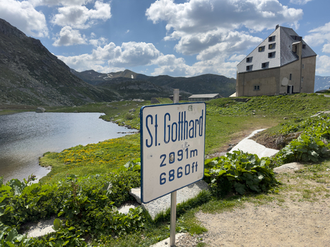 View of the St. Gotthard Pass sign overlooking a mountain lake and a historic stone building under a cloudy sky in Airolo Ticino Switzerland.