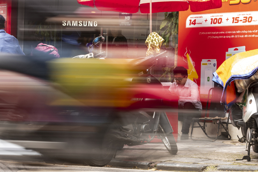 Hanoi Vietnam 11 November 2025 View of a busy street in Hanoi with a motion blurred motorbike passing by and a man sitting on a parked motorcycle near a shop front.