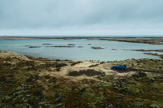 Aerial view of Hlidarvatn lake surrounded by mossy volcanic terrain and rocky landscape with a blue car parked on the shore in Sveitarfelagid Olfus Iceland. Aerial view of Hlidarvatn lake surrounded by mossy volcanic terrain and rocky landscape with a blue car parked on the shore in Sveitarfelagid Olfus Iceland.