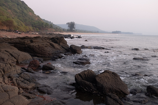View of dark jagged rocks meet the foamy surf under a hazy sky where the shoreline curves towards distant hills Chiplun Maharashtra India. View of dark jagged rocks meet the foamy surf under a hazy sky where the shoreline curves towards distant hills Chiplun Maharashtra India.