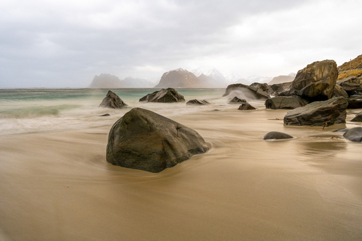 View of large rocks on a sandy beach with long exposure waves and distant snow capped mountains under an overcast sky in Leknes Lofoten Norway.