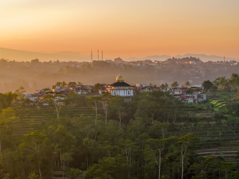 Aerial view of the Dieng Plateau featuring a mosque with a golden dome nestled among lush trees and misty hills under the warm glow of sunrise in Indonesia.