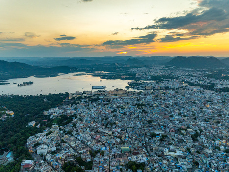 Aerial view of Lake Pichola and the City Palace complex surrounded by dense urban rooftops under a golden sunset sky in Udaipur Rajasthan India.