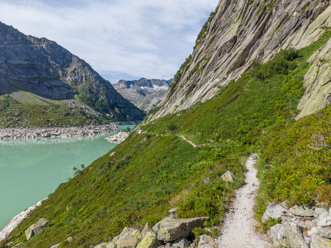 Aerial view of Gelmersee alpine reservoir with a hiking trail along the steep rocky mountainside and turquoise water in Switzerland.