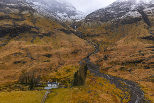 Aerial view of a white cottage at the base of the rugged snow capped mountains in Glencoe Scotland United Kingdom. Aerial view of a white cottage at the base of the rugged snow capped mountains in Glencoe Scotland United Kingdom.