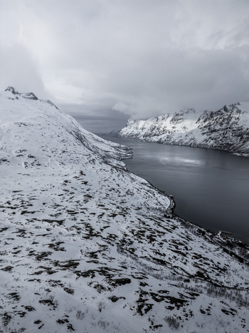 Aerial view of the Ersfjorden fjord surrounded by steep snow covered mountains and dark water under a cloudy winter sky Ersfjord Troms Norway. Aerial view of the Ersfjorden fjord surrounded by steep snow covered mountains and dark water under a cloudy winter sky Ersfjord Troms Norway.