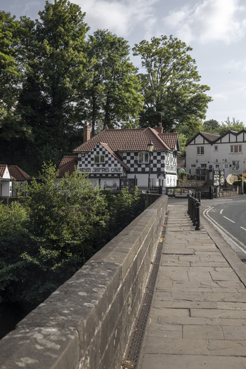 Knaresborough England United Kingdom 14 July 2026 View of Mother Shipton s Cave entrance with its black and white checkered facade lush trees and a stone bridge in the foreground.