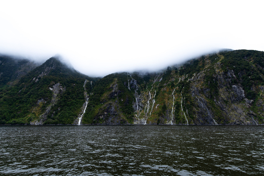View of majestic steep green mountains cascading with slender waterfalls meet the dark rippled waters under a misty overcast sky Milford Sound New Zealand.