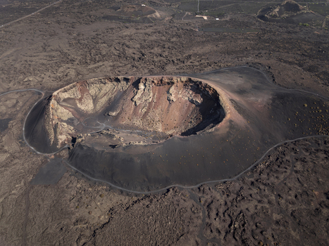 Aerial view of a volcanic crater with rugged dark lava fields reddish rock formations and a winding path around the rim in Lanzarote Canarias Spain. Aerial view of a volcanic crater with rugged dark lava fields reddish rock formations and a winding path around the rim in Lanzarote Canarias Spain.