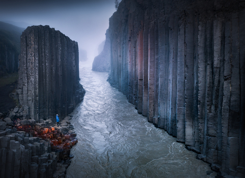 Aerial view of Studlagil Canyon featuring towering basalt columns and a glacial river with a person in a blue jacket standing on the rocks under a misty sky Iceland.