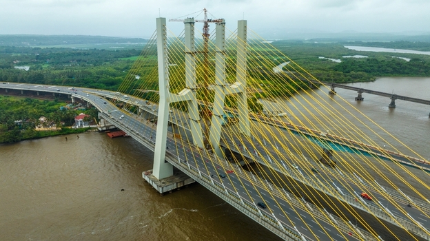 Aerial view of the Zuari Bridge with its cable stayed design yellow cables and construction crane spanning the river with lush green banks and nearby infrastructure in Cortalim Goa India.