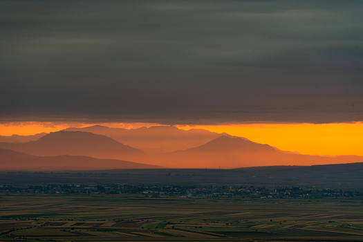 View of a vibrant sunset casting a golden glow over the distant Carpathian Mountains and the rural valley landscape in Mereni Covasna County Romania. View of a vibrant sunset casting a golden glow over the distant Carpathian Mountains and the rural valley landscape in Mereni Covasna County Romania.