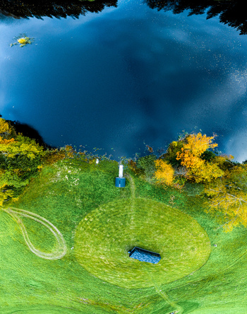 Aerial view of a small structure and a circular path in a lush green field next to a calm blue lake surrounded by autumn trees in Latvia. Aerial view of a small structure and a circular path in a lush green field next to a calm blue lake surrounded by autumn trees in Latvia.
