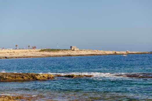 View of azure waters meet the rocky shore under a pale sky with a distant structure standing sentinel over the land Marsaskala Malta. View of azure waters meet the rocky shore under a pale sky with a distant structure standing sentinel over the land Marsaskala Malta.