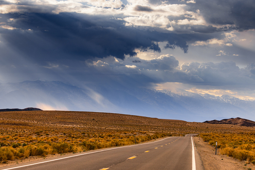 View of Death Valley National Park with a road leading through the desert towards mountains under a dramatic sky with sunbeams breaking through clouds United States.