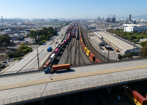 Aerial view of a busy freight railyard with multiple tracks and colorful shipping containers adjacent to a highway with a semi truck driving on an overpass in Carson California United States.
