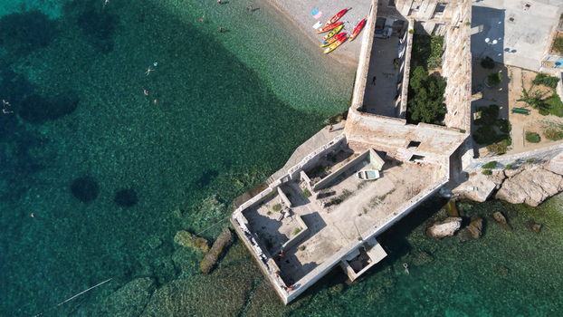 Aerial view of historic stone ruins by the sea with a pebble beach colorful kayaks and people swimming in clear turquoise water in Komiza Split Dalmatia County Croatia.