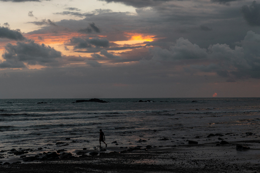 View of a person walking along the wet sandy beach at sunset with dark dramatic clouds over the ocean and scattered rocks in Costa Rica.
