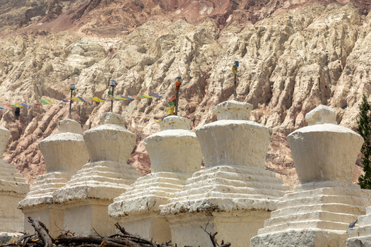 View of white stupas at Charasa Gonpa adorned with colorful prayer flags against a backdrop of rugged mountains in Charasa Ladakh India.