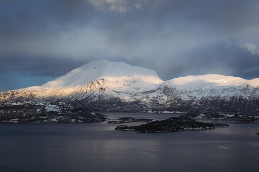 Aerial view of mountains capped with snow reflecting the light of the setting sun over the dark water Sjøvegan Troms Norway. Aerial view of mountains capped with snow reflecting the light of the setting sun over the dark water Sjøvegan Troms Norway.