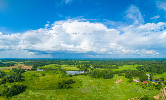 Aerial view of the lush green rural landscape featuring rolling fields dense forests small ponds and a dramatic large cumulus cloud formation in the blue sky Latvia. Aerial view of the lush green rural landscape featuring rolling fields dense forests small ponds and a dramatic large cumulus cloud formation in the blue sky Latvia.
