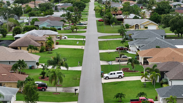 Aerial view of a quiet residential neighborhood with suburban houses lush green lawns palm trees and parked cars along a straight road in Cape Coral Florida United States. Aerial view of a quiet residential neighborhood with suburban houses lush green lawns palm trees and parked cars along a straight road in Cape Coral Florida United States.
