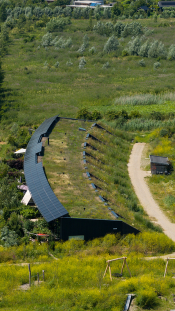 Aerial view of a modern building with a green roof and integrated solar panels set within a lush natural landscape with a dirt path in Almere Flevoland Netherlands. Aerial view of a modern building with a green roof and integrated solar panels set within a lush natural landscape with a dirt path in Almere Flevoland Netherlands.