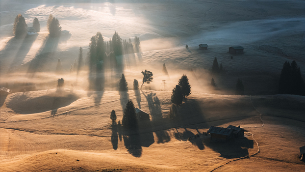 Aerial view of Alpe di Siusi with wooden huts and pine trees shrouded in morning mist and golden light rays Castelrotto Trentino Alto Adige Italy. Aerial view of Alpe di Siusi with wooden huts and pine trees shrouded in morning mist and golden light rays Castelrotto Trentino Alto Adige Italy.