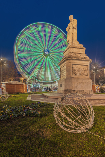 View of the statue of Camillo Benso Count of Cavour illuminated at night with a motion blurred Ferris wheel and festive Christmas decorations in Ancona Marche Italy.