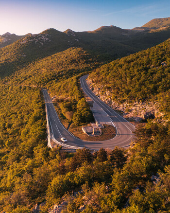 Aerial view of a winding mountain road with a sharp hairpin turn and a car driving through lush green hills at sunset Rijeka Primorje Gorski Kotar Croatia. Aerial view of a winding mountain road with a sharp hairpin turn and a car driving through lush green hills at sunset Rijeka Primorje Gorski Kotar Croatia.