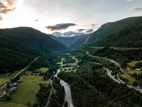 View of Kylling Bridge spanning the Rauma River in a lush green valley with surrounding mountains and winding roads under a soft sunset sky Rauma Municipality More og Romsdal Norway. View of Kylling Bridge spanning the Rauma River in a lush green valley with surrounding mountains and winding roads under a soft sunset sky Rauma Municipality More og Romsdal Norway.