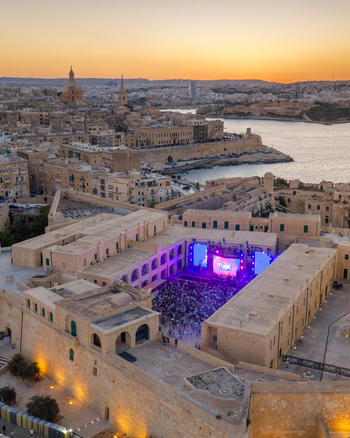 Aerial view of an outdoor concert in a historic stone courtyard with a crowd stage lighting and the city skyline and harbor in the background at sunset Valletta Port Region Malta. Aerial view of an outdoor concert in a historic stone courtyard with a crowd stage lighting and the city skyline and harbor in the background at sunset Valletta Port Region Malta.