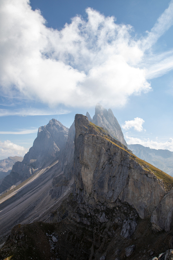 View of the jagged peaks of the Dolomites with steep rocky slopes and a grassy ridge under a blue sky with white clouds in Rocca Pietore Veneto Italy. View of the jagged peaks of the Dolomites with steep rocky slopes and a grassy ridge under a blue sky with white clouds in Rocca Pietore Veneto Italy.