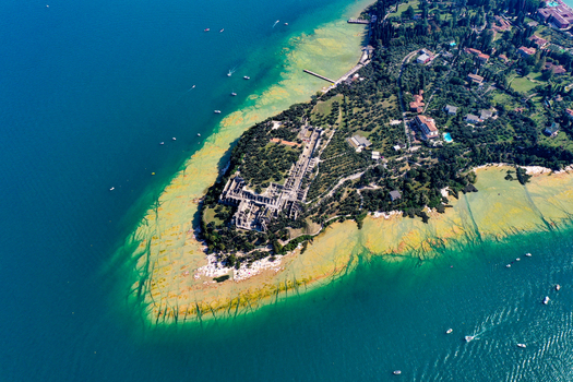 Aerial view of Jamaica Beach and the Grotte di Catullo ruins on the tip of the Sirmione peninsula surrounded by the turquoise waters of Lake Garda Sirmione Lombardia Italy.