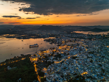 Aerial view of the historic city of Udaipur at sunset featuring the illuminated City Palace and Lake Pichola surrounded by mountains in Udaipur Rajasthan India.