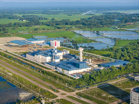 Aerial view of the Mirsarai Economic Zone featuring industrial buildings with blue roofs a chimney and a water tower set among green fields in Minsarai Chittagong Division Bangladesh. Aerial view of the Mirsarai Economic Zone featuring industrial buildings with blue roofs a chimney and a water tower set among green fields in Minsarai Chittagong Division Bangladesh.