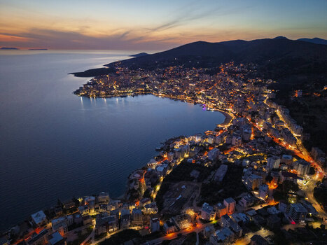 Aerial view of Saranda illuminated at twilight along the Ionian Sea featuring dense urban architecture glowing street lights and the calm bay in Saranda Vlore County Albania. Aerial view of Saranda illuminated at twilight along the Ionian Sea featuring dense urban architecture glowing street lights and the calm bay in Saranda Vlore County Albania.
