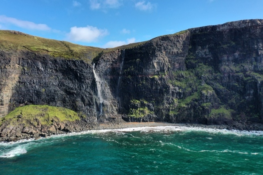 Aerial view of Neist Point waterfall cascading down rugged dark cliffs into the turquoise sea with green grassy slopes in Isle of Skye Scotland United Kingdom. Aerial view of Neist Point waterfall cascading down rugged dark cliffs into the turquoise sea with green grassy slopes in Isle of Skye Scotland United Kingdom.