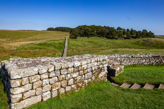 View of the ancient stone ruins of Vindolanda Roman fort with rolling green hills and a clear blue sky in Hexham England United Kingdom. View of the ancient stone ruins of Vindolanda Roman fort with rolling green hills and a clear blue sky in Hexham England United Kingdom.
