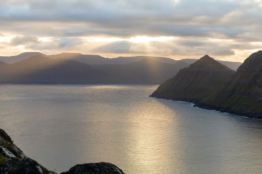 View of the dramatic landscape of the Faroe Islands with steep green mountains rising from the sea under a golden sunset sky with sunbeams breaking through clouds Faroe Islands. View of the dramatic landscape of the Faroe Islands with steep green mountains rising from the sea under a golden sunset sky with sunbeams breaking through clouds Faroe Islands.