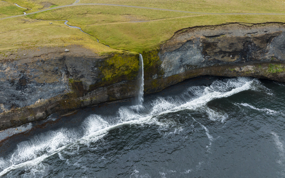Aerial view of a waterfall cascading down a rugged cliff into the turbulent sea the frothy waves crashing against the dark rocks below H&uacute;nabygg&eth; Iceland.