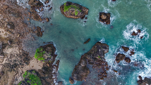 Aerial view of a rocky coastline with turquoise water crashing waves and lush green vegetation on dark stone formations along the shore of Indonesia. Aerial view of a rocky coastline with turquoise water crashing waves and lush green vegetation on dark stone formations along the shore of Indonesia.