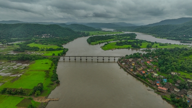 Aerial view of the Mumbai Goa Highway bridge spanning a wide river surrounded by lush green fields rolling hills and a small village in Mahad Maharashtra India.