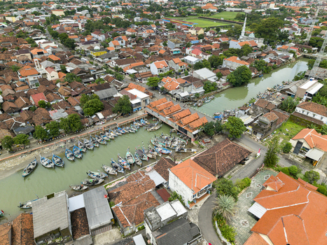 Aerial view of fishing boats docked along the murky riverbanks beneath the bridge contrasting with the surrounding red tiled roofs Jepara Central Java Indonesia.