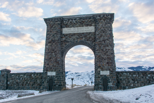 View of the historic stone Roosevelt Arch at the north entrance of Yellowstone National Park set against a snowy mountain landscape under a cloudy sky in the United States.