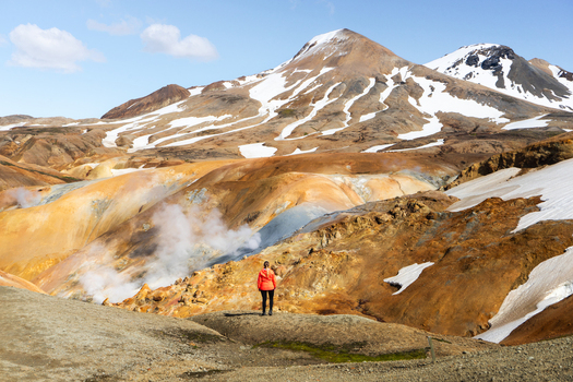 View of a hiker in an orange jacket standing in the geothermal landscape of Kerlingarfjoll with steaming vents colorful rhyolite mountains and patches of snow in Hrunamannahreppur Iceland.
