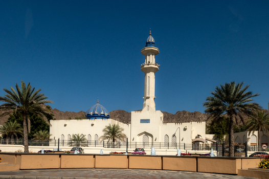 View of a mosque with a prominent minaret and blue dome surrounded by palm trees and mountains under a clear blue sky in Muscat Muscat Governorate Oman. View of a mosque with a prominent minaret and blue dome surrounded by palm trees and mountains under a clear blue sky in Muscat Muscat Governorate Oman.